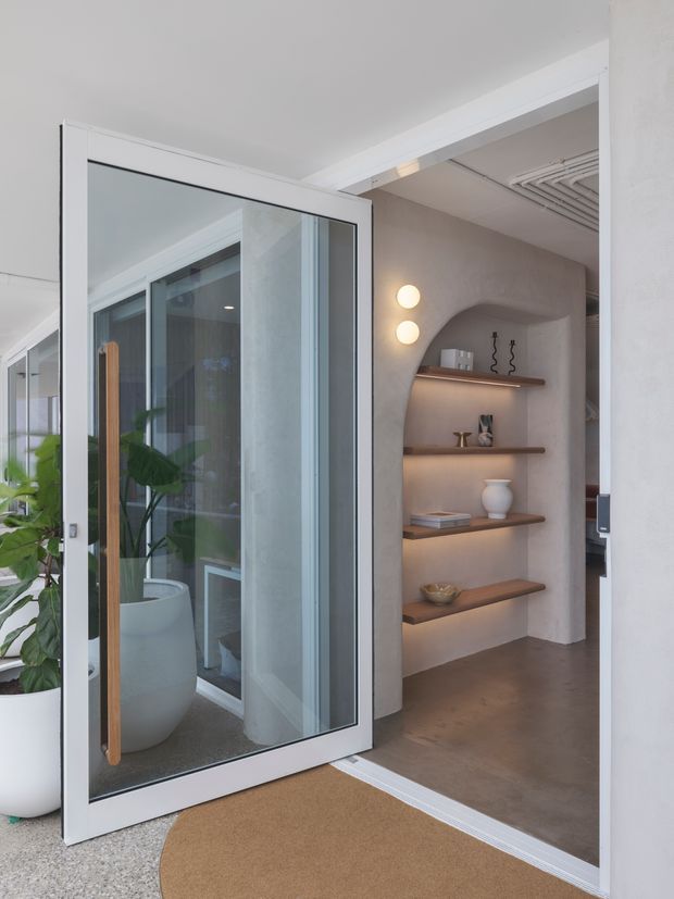 A bright, minimalist residential entryway viewed through an open glazed door. The space features white walls and ceiling, a large white-framed glass pivot door with a timber pull handle, a terrazzo-like light floor at the threshold, and a warm brown interior floor. Inside, a curved plaster niche contains three floating timber shelves with integrated warm lighting and understated decorative objects. A large potted plant is visible through the glazing, and a linear ceiling vent appears deeper in the adjoining space.