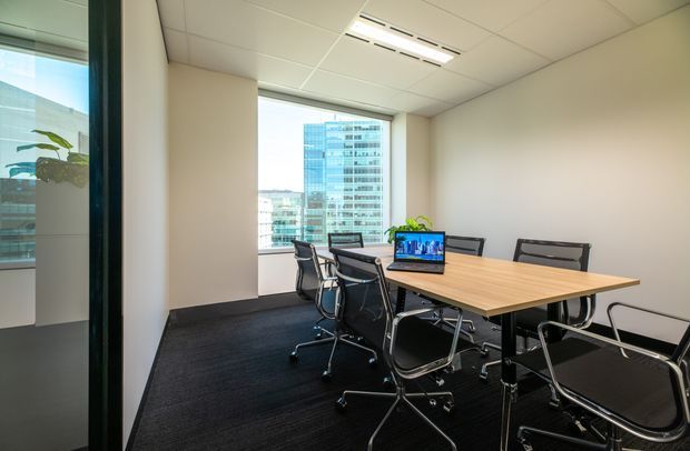 A bright, modern commercial meeting room with a rectangular light-wood conference table, black mesh office chairs, carpeted flooring, white walls, a suspended tiled ceiling with integrated linear air-conditioning, and a large window overlooking an urban office building. A glass partition and reflected greenery are visible at the left.