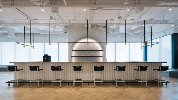 A bright contemporary commercial hospitality or breakout space with a long fluted-front counter and evenly spaced black bar stools. The room features a large arched illuminated wall niche or shelving feature, extensive floor-to-ceiling glazing, exposed white services and ductwork, suspended linear pendant lighting, and a light timber parquet floor. The restrained palette of white, pale timber, black, and blue creates a clean, airy, modern workplace or venue interior.