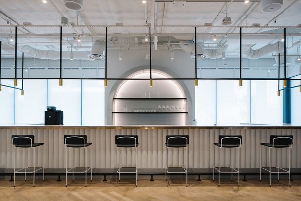 A bright, contemporary commercial hospitality space with a long light-wood bar counter and vertically fluted front, lined with minimalist black bar stools. Behind the counter is an illuminated arched drinks display, while full-height windows, black-framed glazed partitions, exposed white services, ductwork and suspended pendant lights create an open-plan cafe or bar atmosphere.