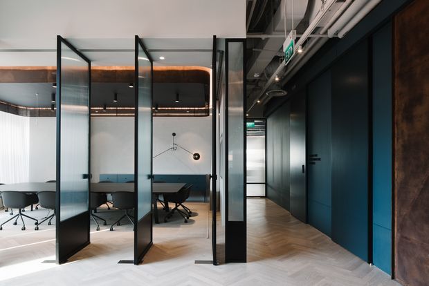 A contemporary commercial office interior with glazed, black-framed meeting-room partitions on the left and a dark corridor with exposed services on the right. The meeting room contains a long table, office chairs, and a blue upholstered banquette. Herringbone timber flooring, dark blue-black walls and joinery, translucent glass panels, exposed ceiling pipes and ducts, and warm linear lighting create a refined industrial workplace atmosphere.