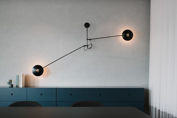 A refined dining area with a long dark tabletop in the foreground, a low built-in dark teal sideboard spanning the wall, and a sculptural black wall-mounted light fixture with three articulated arms and warm glowing bulbs. The space has a neutral grey wall, sheer curtains at the right, and restrained contemporary styling.