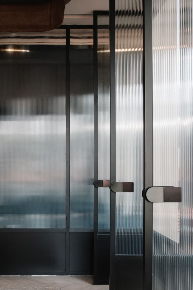 Close-up interior view of a dark-framed glazed partition or set of doors with vertically ribbed translucent glass panels. Repeated black metal mullions and rounded two-tone handles create a refined industrial look, with a herringbone timber floor and a dark wood element visible at the top left.