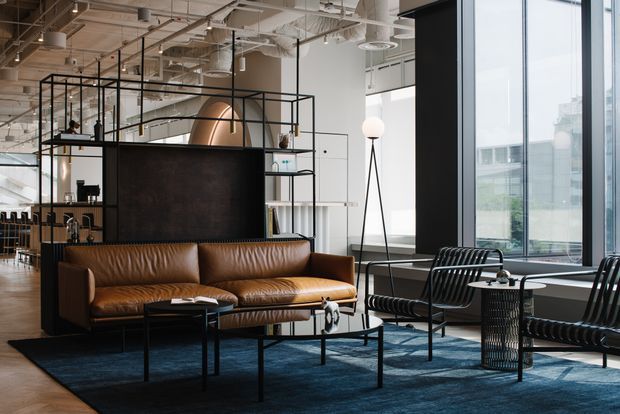 A contemporary open-plan commercial lounge or reception area with floor-to-ceiling windows, exposed services and ceiling structure, black metal-framed shelving, a dark partition, and leather seating arranged around low coffee tables. The space has an industrial-modern character with warm timber and brown leather balanced by dark metal, concrete-grey finishes, and abundant daylight.