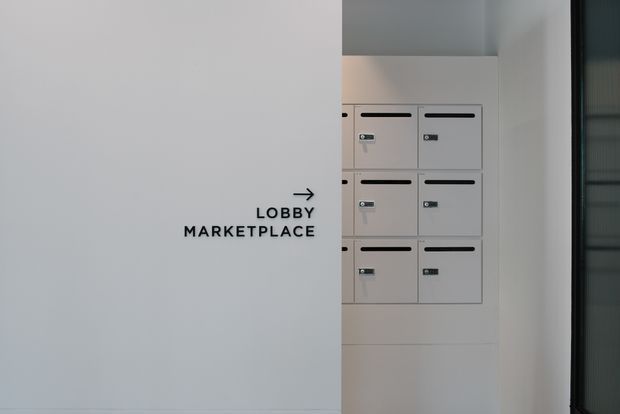 Minimalist interior signage directing visitors toward a lobby marketplace, with a wall-mounted grid of white mailboxes and a dark-framed glazed opening at right. The space is clean, neutral, and institutional or commercial in character.