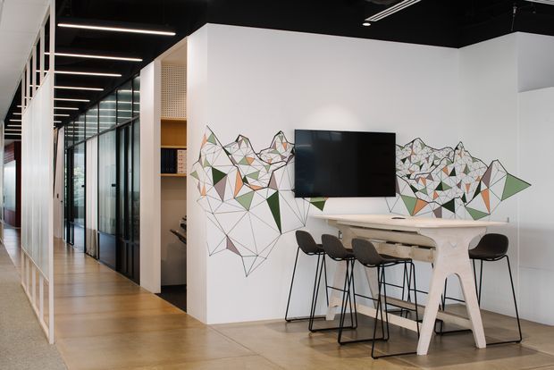 A contemporary commercial-style interior with an open breakout or meeting area beside a glazed corridor. The space features a light timber floor, white walls, black exposed ceiling, linear strip lighting, black-framed glass partitions, a wall-mounted television, and a high white communal table with black bar stools. A geometric abstract mural in muted green, terracotta, grey, and black spans the feature wall.