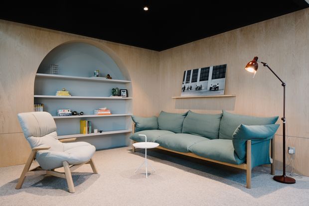 A softly lit contemporary living room with pale timber wall panelling, a black ceiling, and a built-in arched shelving niche displaying books and small objects. The room contains a muted teal sectional sofa, matching lounge chair, small white side table, framed wall art, and an adjustable floor lamp, creating a calm, refined setting.