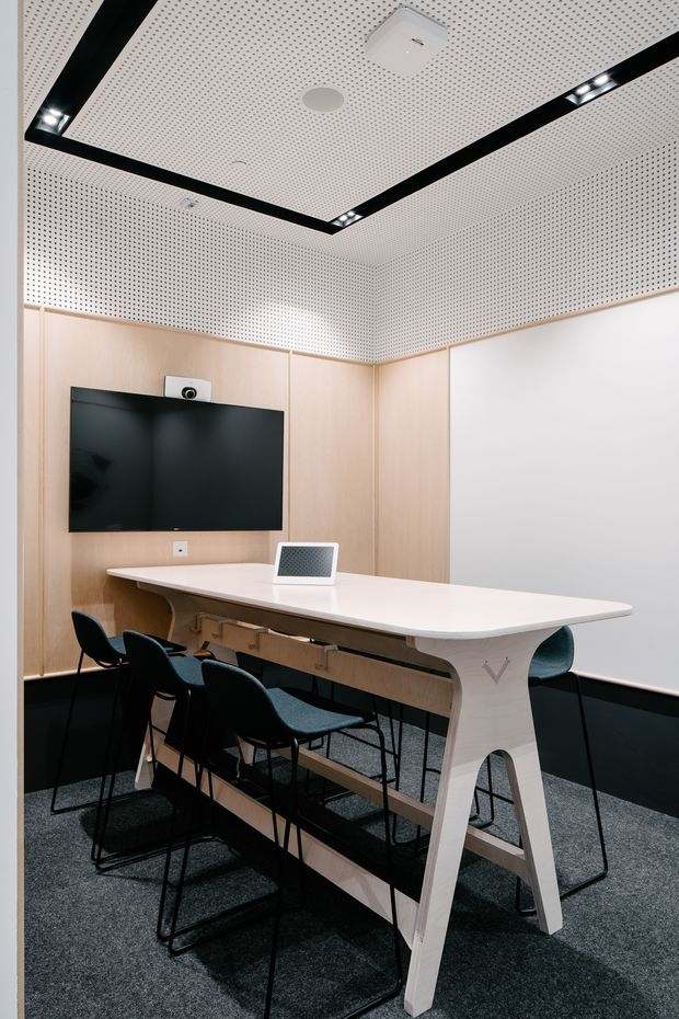 A compact contemporary meeting room or video-conferencing room with pale timber wall panelling, a large wall-mounted display and camera, a white integrated conference table, and dark upholstered chairs. The room has a dark carpeted floor, acoustic perforated wall and ceiling panels, and recessed black linear lighting.