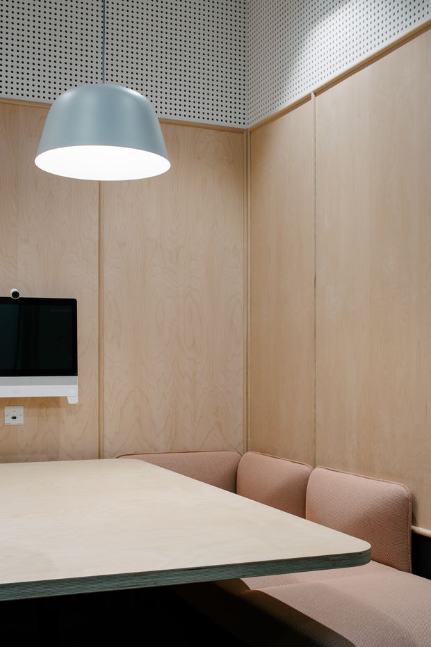 Compact contemporary meeting or dining room with pale plywood wall panelling and built-in upholstered bench seating around a light timber table. A perforated acoustic ceiling and matching wall treatment add texture, while a large matte grey pendant provides focused illumination. A wall-mounted screen and power outlet suggest a small office meeting space or collaborative workspace.