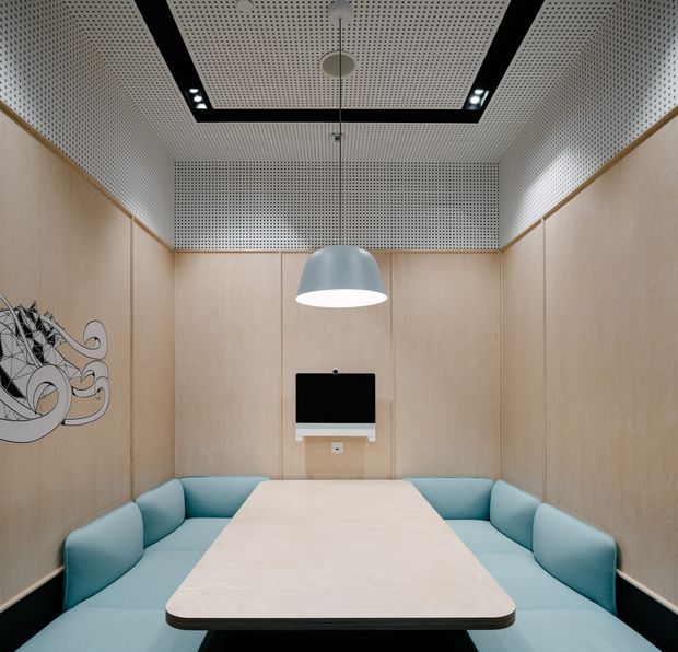 Compact contemporary meeting booth with pale timber wall panelling, integrated light-blue upholstered bench seating, and a central pale timber table. The room features a perforated acoustic ceiling and rear wall, recessed spotlights, a suspended pendant light, a wall-mounted display, and a small monochrome graphic at the left edge.