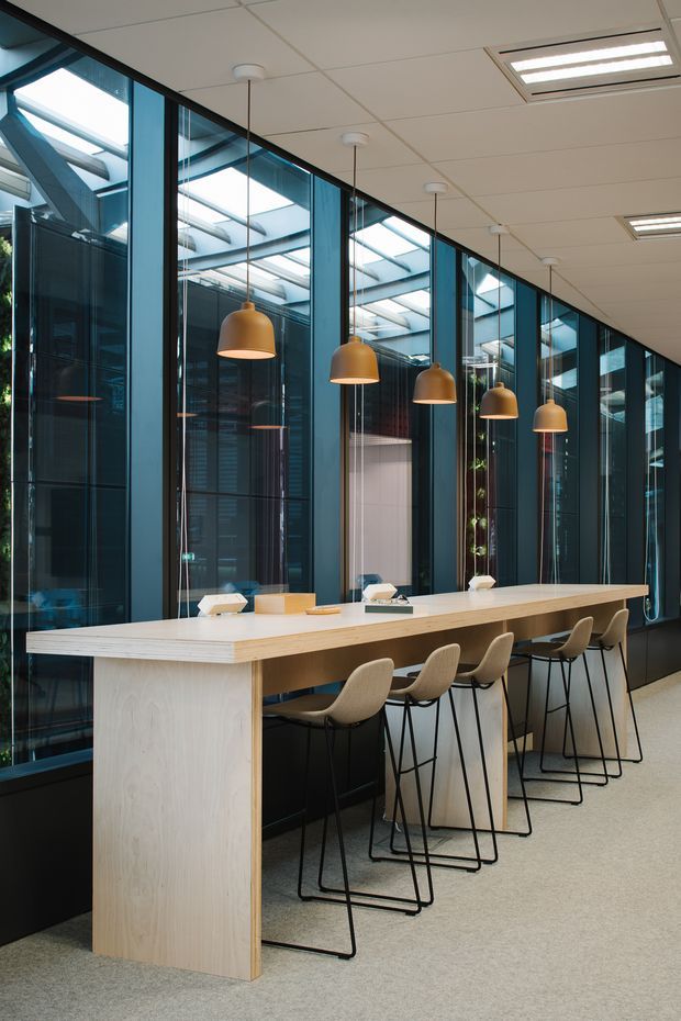 A contemporary commercial workspace with a long pale timber communal counter and integrated seating. The counter runs alongside a dramatic wall of full-height black-framed glazing, revealing a glazed atrium or internal lightwell with skylights and structural steel beams. Warm pendant lights hang above the counter, while recessed fluorescent-style ceiling panels provide general illumination. The space has carpet flooring, dark glazing frames, and a refined, functional office fit-out.