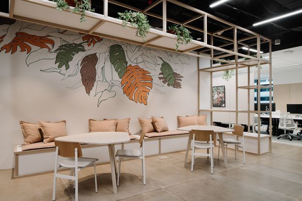 A contemporary commercial office breakout and dining area with built-in banquette seating, round tables, timber chairs, and a large botanical mural. Open timber-framed screening and overhead planter shelves divide the space from a visible open-plan workstation area. The interior has a polished concrete floor, light neutral walls and joinery, and a black exposed ceiling with linear lighting.