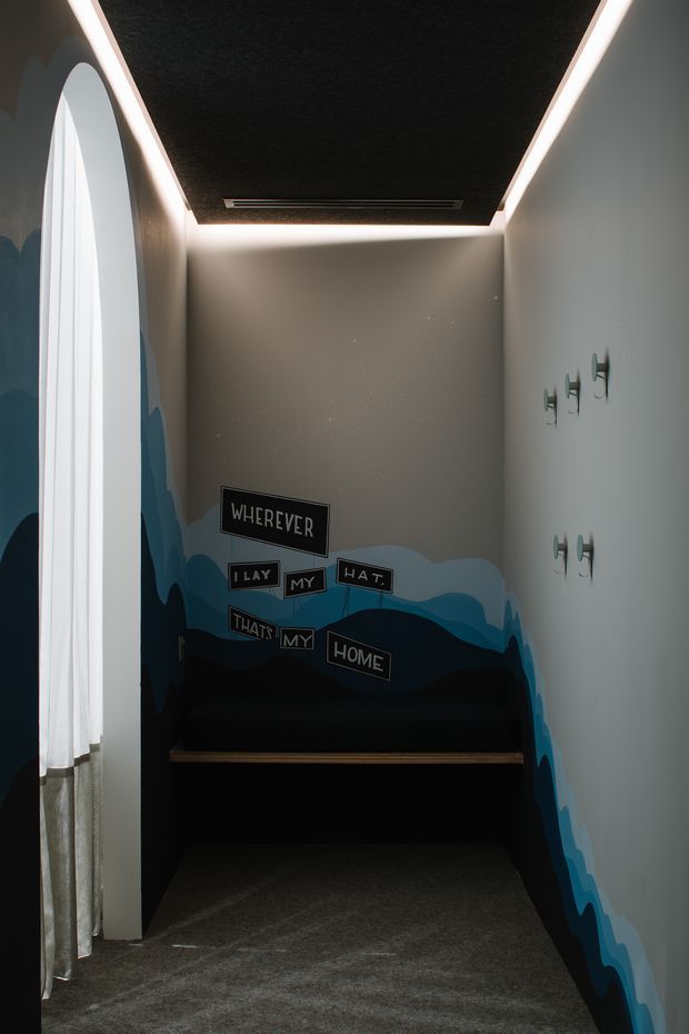A narrow, moody interior alcove or hallway with dark carpet, grey walls, a black textured ceiling panel edged with concealed linear lighting, and a deep built-in bench or storage unit at the end. A tall arched window with sheer curtains occupies the left side, while a layered blue wave-like mural wraps around the lower walls. Small sculptural hooks are mounted on the right wall, and playful typographic wall plaques read “WHEREVER I LAY MY HAT, THAT’S MY HOME.”