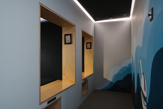 A narrow contemporary hallway or corridor with integrated timber-lined recessed display niches and lower storage compartments. The walls are painted in cool grey-blue tones, with a large blue and navy abstract mural along the right wall. Recessed linear lighting traces the ceiling edges, and a dark ceiling panel adds contrast. The space has a dark carpeted floor and a clean, modern commercial or institutional fit-out.