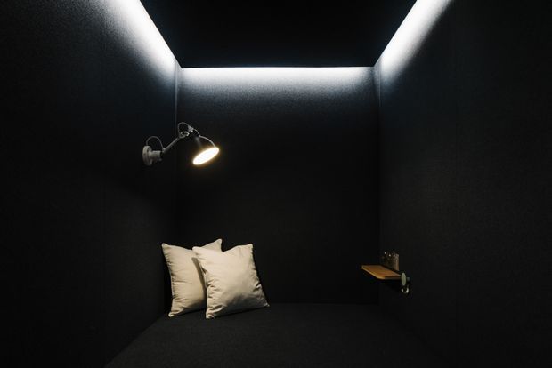 A compact, very dark monochromatic room with black wall, ceiling, and built-in daybed surfaces. Two cream pillows sit at the back, while a small wall-mounted spotlight provides a warm pool of light. A narrow shelf with a few small objects is mounted on the right wall, and concealed linear lighting outlines the recessed ceiling edge.
