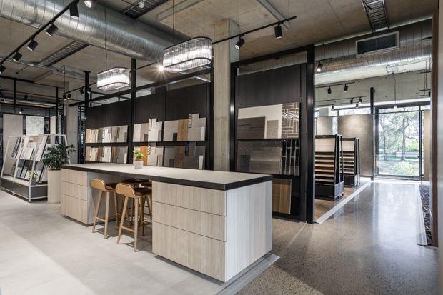 Contemporary flooring and materials showroom with a central light-wood display island and stools, extensive sample boards, polished concrete flooring, exposed concrete columns, black steel framing, exposed ductwork, track lighting, and large glazed frontage opening to greenery outside.