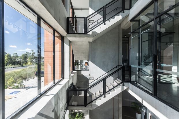 A bright multi-level interior atrium with exposed concrete walls and stair flights zigzagging around a central void. Black metal railings, extensive floor-to-ceiling glazing, and timber-framed vertical window panels create a modern, industrial architectural character. Reflections reveal additional glazing and interior circulation spaces, with landscaped exterior views visible beyond the windows.