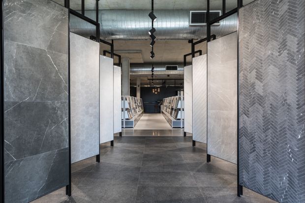 A modern commercial showroom interior viewed through a corridor of freestanding tile display panels. Large-format dark grey stone-look floor and wall tiles frame the space, with patterned herringbone and geometric tile surfaces on the display boards. The ceiling has exposed concrete, ductwork, black metal framing, and linear pendant lighting, creating an industrial retail showroom atmosphere.
