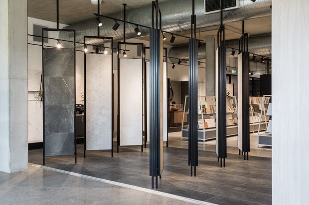A contemporary commercial showroom or design studio interior with freestanding black metal-framed display partitions, stone and tile sample boards, dark flooring, concrete structural elements, exposed services, and track lighting. The space has an industrial, curated materials-library atmosphere.