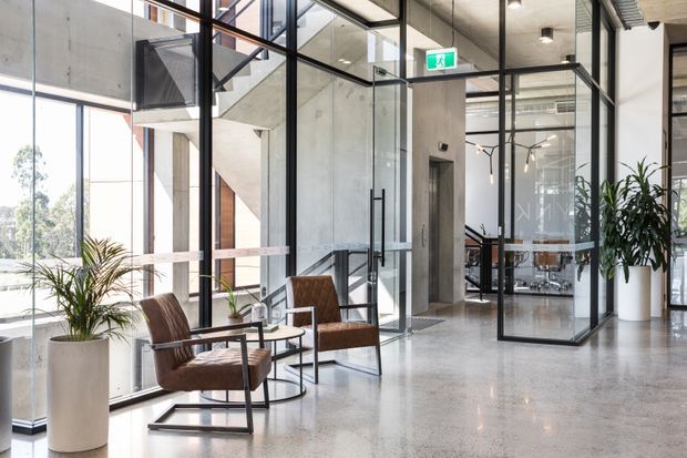 Bright contemporary commercial interior with a polished concrete floor, exposed concrete structural elements, extensive black-framed glass partitions, and a glazed meeting room. A small waiting area with two leather lounge chairs and side tables sits beside floor-to-ceiling windows, with indoor plants adding greenery. The space has an airy, refined office or commercial lobby character.