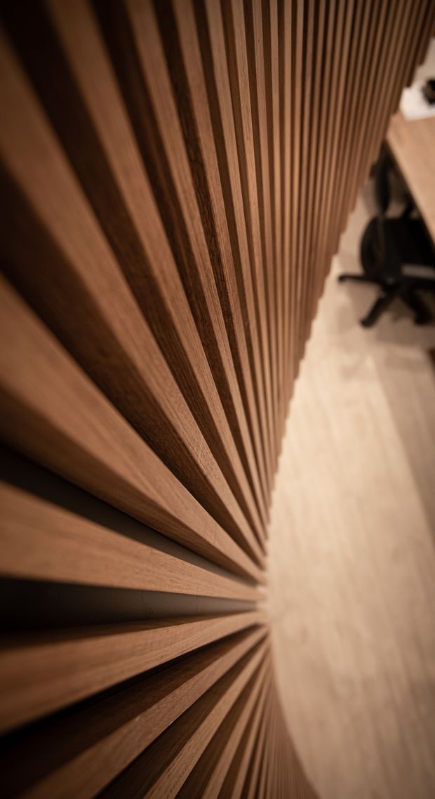 Close-up interior view of a dramatic vertical timber slat feature wall or screen, with deep rhythmic recesses creating strong perspective and shadow lines. A pale timber floor, desk edge, and office chair are visible in the blurred background, suggesting a warm contemporary workspace.