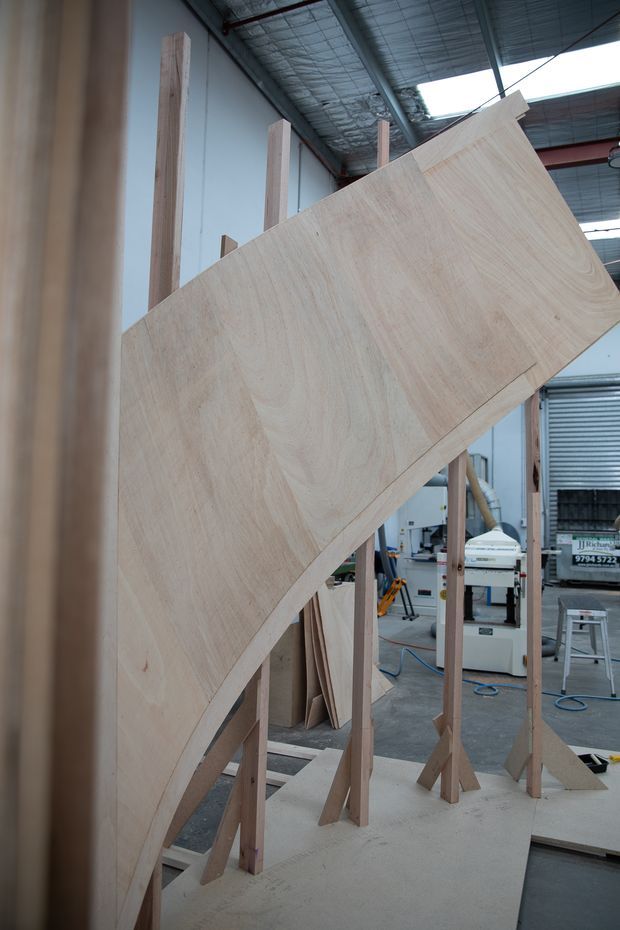A large curved plywood panel is being assembled and supported on timber framing inside a workshop. The industrial workspace includes exposed metal roofing, fabrication machinery, timber supports, and construction materials.
