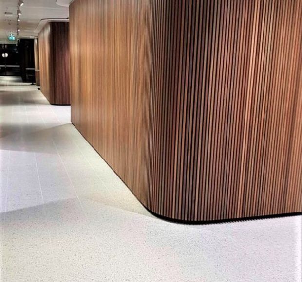 A contemporary interior corridor with gently curved walls clad in vertical warm timber battens. The corridor has a pale speckled hard floor, recessed and track lighting, and dark openings or doors at the far end. The image focuses on the sculptural timber wall treatment and clean circulation space.