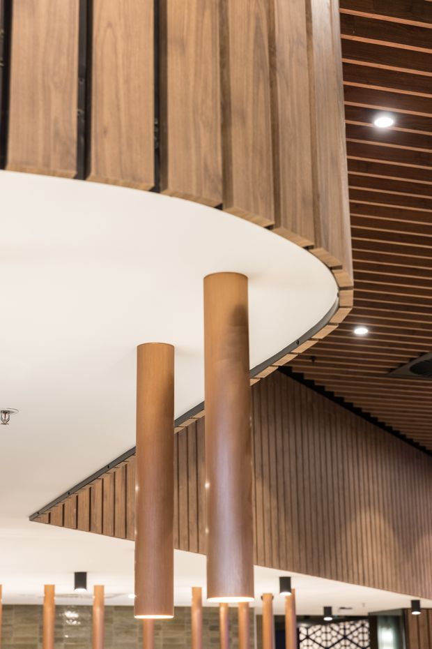 Interior architectural view of a large, refined public or commercial space featuring curved white ceiling forms, tall cylindrical timber-clad columns, extensive vertical timber wall panelling, and a dramatic slatted timber ceiling with recessed lighting. The image emphasizes the sculptural ceiling geometry and warm wood detailing.