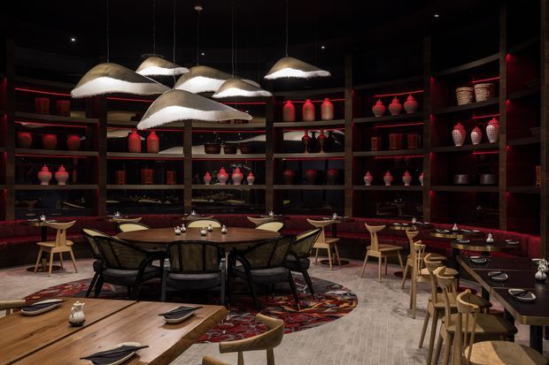 A dark, atmospheric restaurant dining room with extensive dark timber shelving displaying ceramic vessels, red accent lighting, upholstered banquette seating, and multiple communal and intimate dining tables. Sculptural pendant lights hang above the central seating area, while patterned rugs, timber tables, and light wood chairs add warmth and visual contrast.
