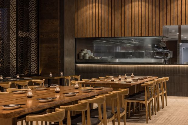 A warmly lit contemporary restaurant dining room with long timber tables, wooden chairs, ceramic tableware, and pendant-like table lamps. A visible open kitchen is framed by dark tile and timber wall panelling, while decorative patterned wall coverings and a tiled floor add texture.