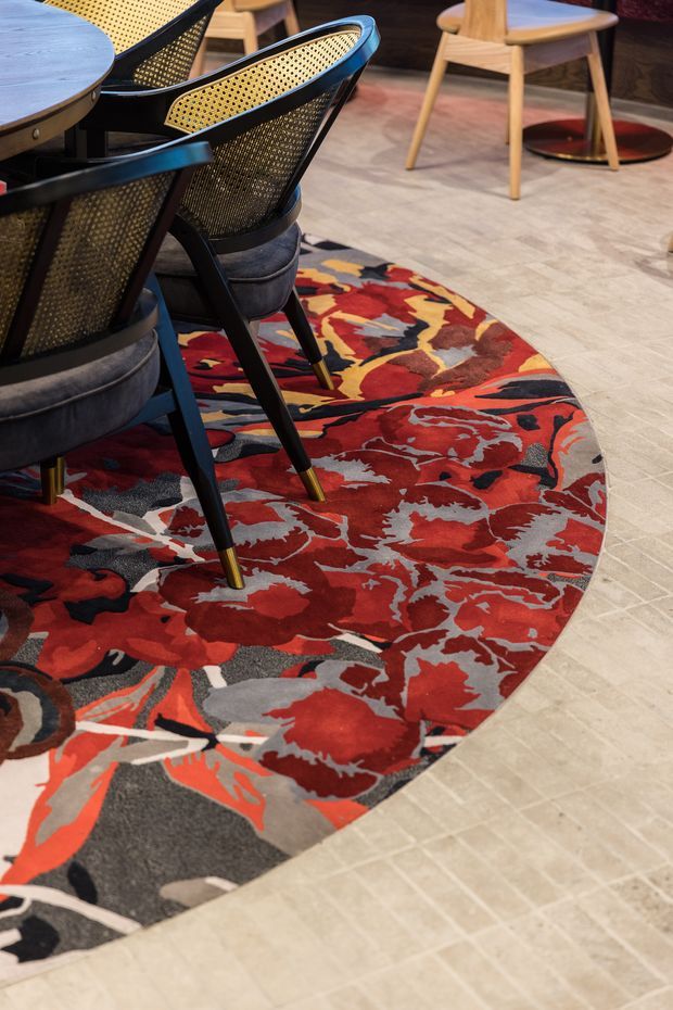 Close-up interior view of a dining area featuring a round wood table and black-framed woven cane dining chairs with upholstered seats. A large, vividly patterned red, charcoal, cream and orange area rug defines the dining zone over a light beige tiled floor. Additional timber seating and a dark pedestal table are visible in the background.