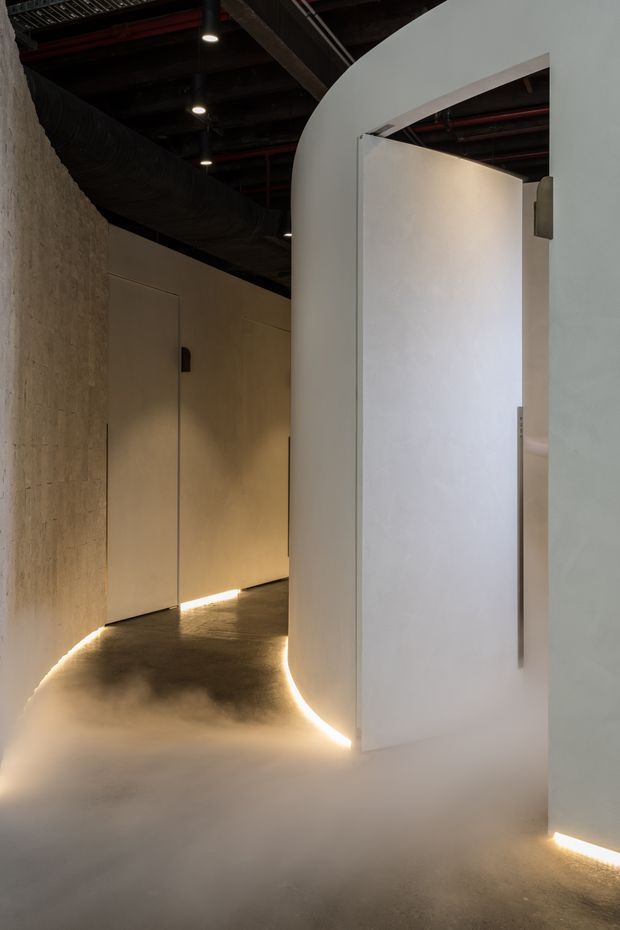 A dimly lit contemporary interior corridor with curved white plaster partitions and concealed doors. Warm linear floor-level lighting traces the walls, contrasting with a dark exposed industrial ceiling featuring ducts, pipes, and spotlights. The floor appears smooth and continuous, with a softly textured concrete-like finish.