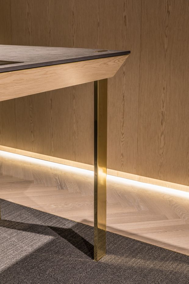 Close-up detail of a refined built-in or freestanding desk/work surface with a light timber top and matching timber wall panelling. A slim brass-gold metal support leg, integrated warm LED strip lighting, pale timber flooring, and dark grey carpet create a warm, contemporary interior composition.