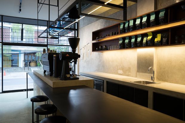 Contemporary commercial café interior featuring a long dark counter with stools, a coffee machine and grinder, a pale stone or composite worktop, black cabinetry, an integrated sink, and illuminated open shelving displaying coffee equipment and supplies. Full-height glazed doors open to an outdoor courtyard or service area, while exposed concrete-look walls, black metal framing, and linear lighting create an industrial atmosphere.