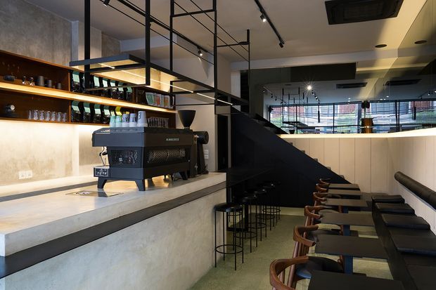 A contemporary café or bar interior with a long concrete service counter, professional espresso machine, illuminated timber shelving displaying bottles and glassware, and a row of stools along a lowered seating area. Black metal-framed suspended shelving and exposed structural elements create a strong industrial character, while track lighting and warm concealed illumination add a refined atmosphere.