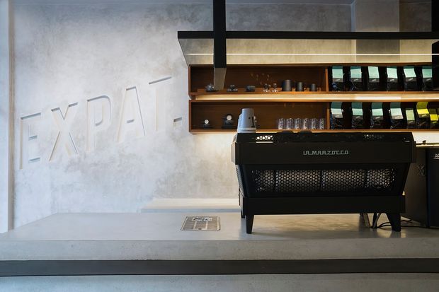 A minimalist coffee bar interior featuring a large black commercial espresso machine on a pale concrete counter. Behind it, dark timber shelving displays cups, glasses, coffee equipment and products, with a textured light grey wall bearing large subtle raised lettering. Black metal framing and warm under-shelf lighting create a refined industrial atmosphere.