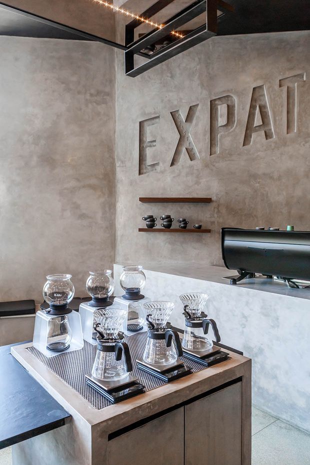 A compact specialty coffee bar with a concrete-finished counter and walls, multiple manual pour-over coffee stations, grinders, and a large black espresso machine. The space features dark exposed ceiling elements, linear pendant lighting, floating timber shelves with coffee vessels, and large dimensional “EXPAT” lettering on the wall. The overall aesthetic is industrial and contemporary.
