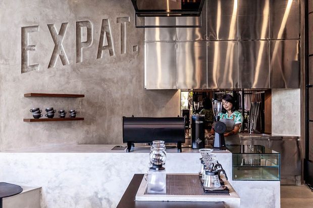 Industrial-style café or coffee bar interior with a polished concrete service counter, stainless-steel ventilation hood, exposed concrete walls, dark metal framing, open shelving with cups, and a barista workstation. Large metallic lettering decorates the feature wall, while reflective glass and stainless-steel surfaces add a contemporary commercial feel.