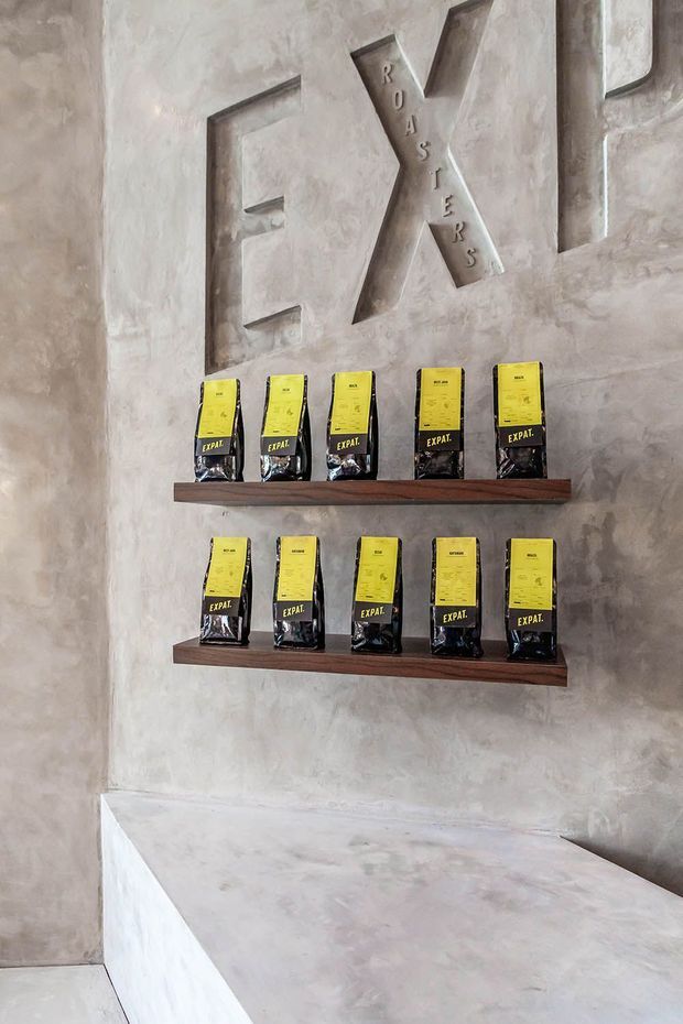 A minimalist retail or hospitality display wall with textured grey plaster/concrete-effect surfaces, recessed geometric lettering, and two dark timber shelves holding rows of yellow-and-black packaged products. A large built-in pale concrete/plaster counter or bench occupies the foreground.