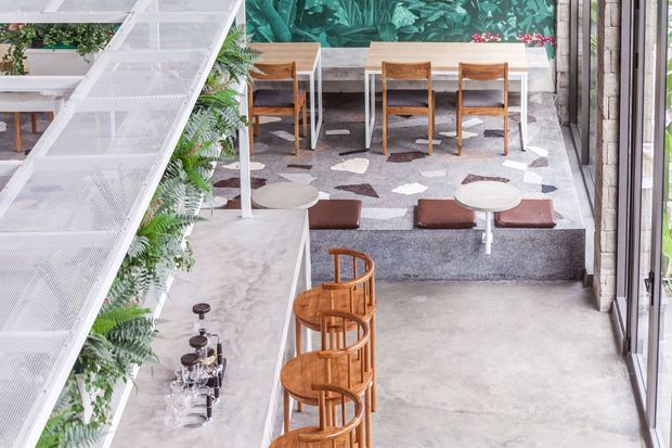 A bright contemporary commercial hospitality interior viewed from above, featuring a long marble-look bar with timber stools, a polished concrete floor, raised dining platforms with timber tables and chairs, and a large botanical mural. A prominent white metal mesh stair or mezzanine structure with integrated greenery occupies the left side.