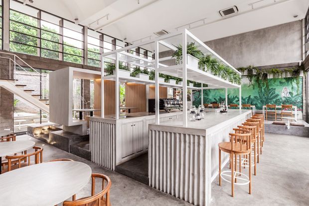 Bright, spacious contemporary commercial café or hospitality interior with a large central bar island, white ribbed cabinetry, timber bar stools, indoor planting, exposed concrete surfaces, extensive black-framed glazing, and a sculptural open stair. A vivid botanical mural and greenery form a feature backdrop.