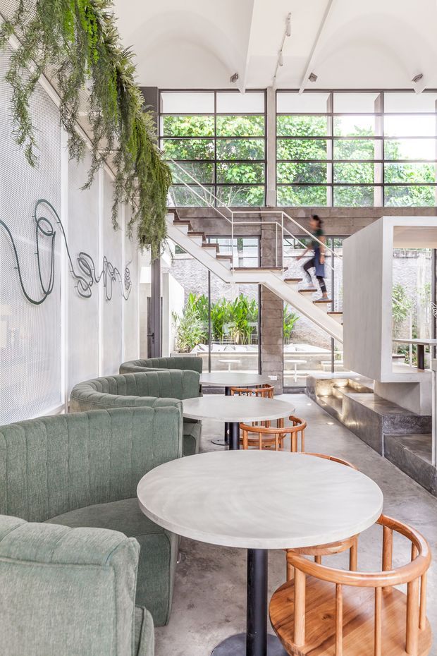 Bright, multi-level contemporary interior featuring a café or hospitality seating area with round tables, curved green upholstered lounge chairs, and timber-framed chairs. A sculptural open stair with slim metal balustrades rises beside a substantial concrete or stone feature wall, while large black-framed industrial-style windows bring in abundant greenery and daylight. Hanging plants and wall artwork add an organic decorative element.