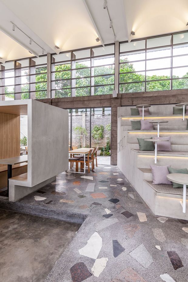Bright contemporary commercial interior with a lofty ceiling, exposed concrete structural beams, extensive gridded windows, built-in stepped upholstered seating, and integrated warm LED lighting. The space features polished concrete flooring and a terrazzo-like patterned carpet or flooring in muted grey, cream, terracotta, and dark tones. Timber-lined booth seating and dining tables are visible, with greenery outside the glazing.