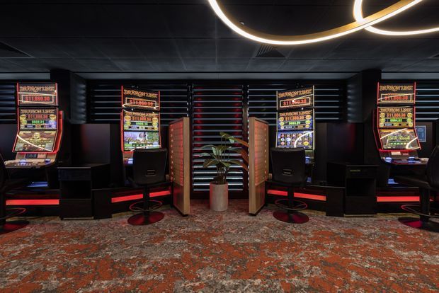 A dark, modern gaming or casino interior featuring multiple illuminated slot machines arranged along a black wall with horizontal slatted detailing. Black gaming chairs, red accent lighting, a central potted plant, sculptural circular ceiling lighting, and a patterned red-and-grey carpet create a dramatic atmosphere.
