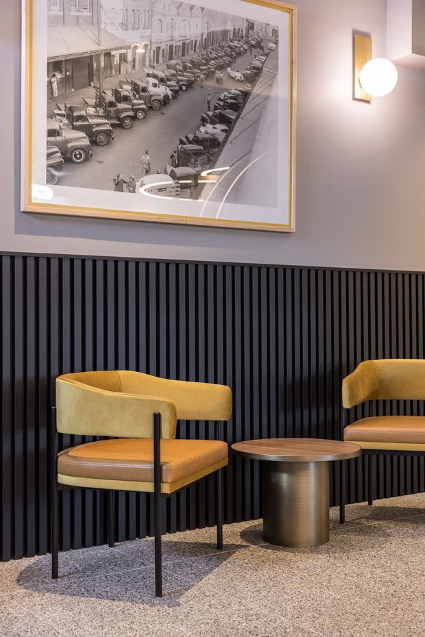 A refined commercial lounge or reception seating area with two mustard upholstered armchairs, a round metallic pedestal coffee table, and a grey textured carpet. The lower wall is clad in black vertical timber slats, contrasting with a soft grey upper wall. A large framed black-and-white historic streetscape photograph and a minimalist wall sconce add visual character. The space has a warm, sophisticated hospitality or lobby feel.