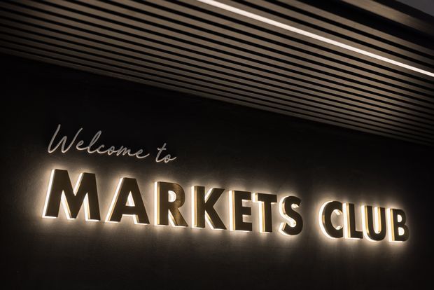 A dark commercial interior featuring a backlit “Welcome to MARKETS CLUB” sign mounted on a textured black wall. The ceiling has closely spaced linear timber-toned slats with recessed strip lighting, creating a dramatic and refined entry or hospitality atmosphere.