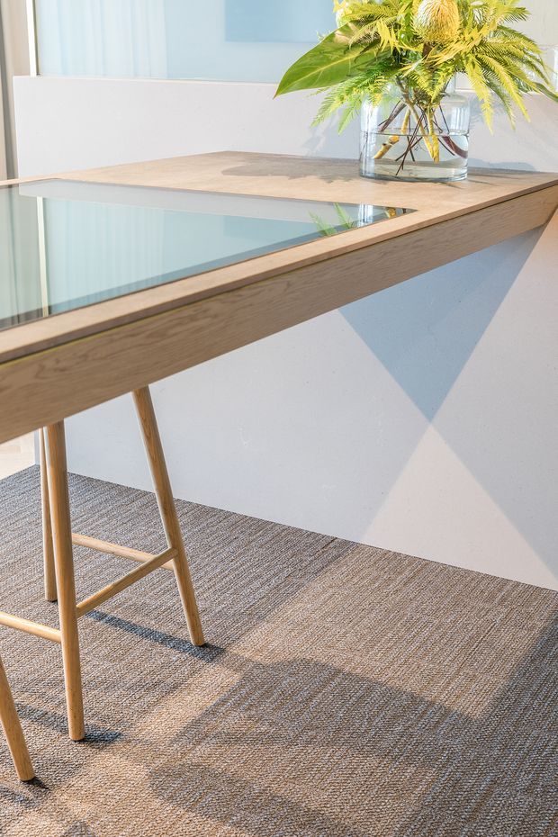 Close-up interior view of a light timber console or desk with a glass inset top, slender angled timber legs, and a potted fern in a clear glass vessel. The piece stands against a pale wall with geometric sunlight and shadow patterns, above a textured neutral carpet.