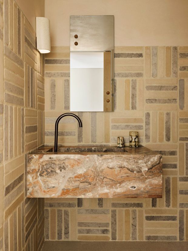 A compact bathroom vanity area with a wall-mounted rectangular stone basin in richly veined warm-toned stone, set against patterned masonry-style wall tiling. A black gooseneck tap, frameless mirrored cabinet, and cylindrical wall light complete the refined, textural composition.