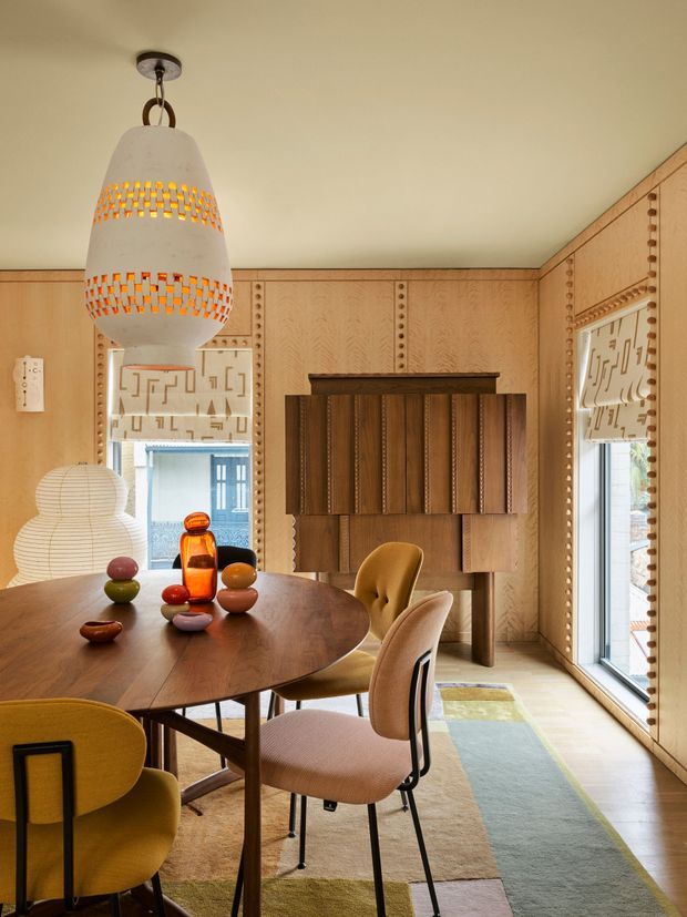A warm, richly detailed dining room with extensive light timber wall panelling and decorative beaded trim. A large round dark-wood dining table is surrounded by upholstered mustard, ochre and tan chairs, with sculptural coloured glass objects as a centrepiece. A tall woven pendant light hangs above the table, while patterned roller blinds, a built-in dark timber cabinet, and a long muted multicolour rug add visual interest. The room has generous windows and a cosy, curated mid-century-inspired atmosphere.