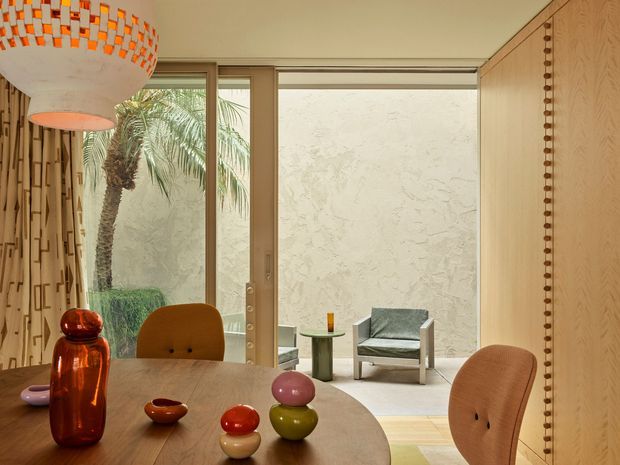 Warm, mid-century-inspired dining area with a round timber dining table, sculptural upholstered chairs, colourful ceramic objects, and a large woven pendant light. Full-height timber-framed glazing opens the room visually to a small outdoor courtyard with a palm, while a pale timber feature wall and decorative vertical detailing frame the space. A separate lounge chair is visible beyond the glazing.